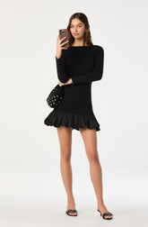 A woman is wearing a fitted black long-sleeve dress with a ribbed texture and a ruffled hemline. She has a small black handbag with white polka dots slung over her shoulder and is holding a smartphone in front of her face. She is also wearing black open-toe heels with decorative embellishments.